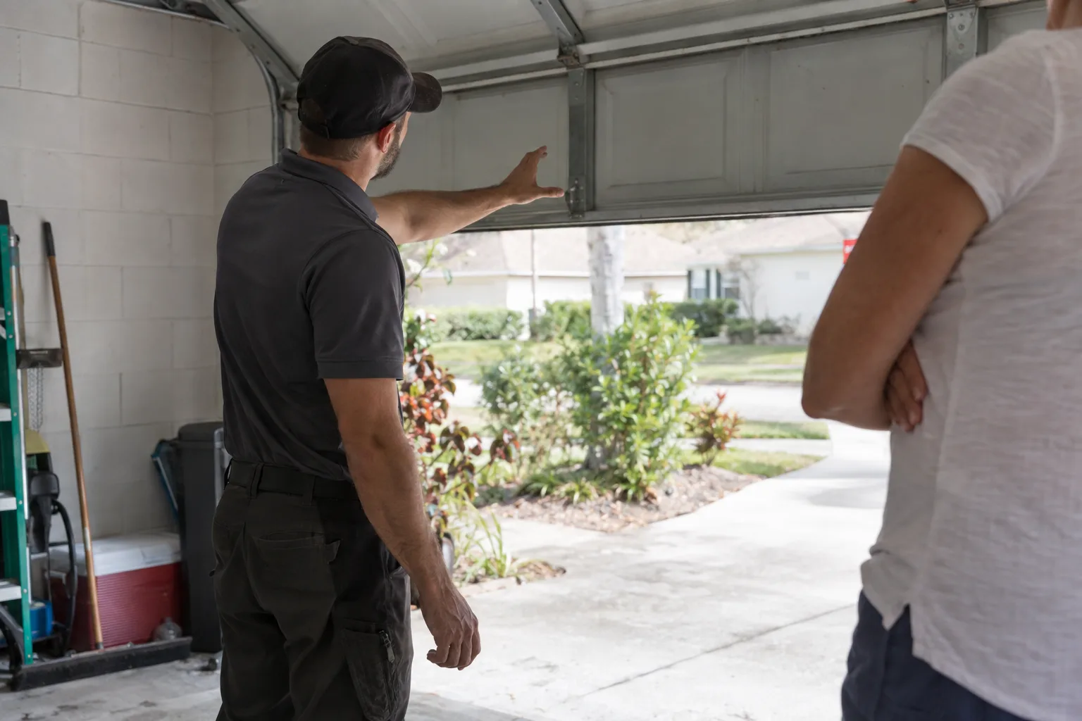 A garage door technician seen from behind gestures toward an open residential garage door panel while a homeowner stands nearby listening, daylight filling a Florida suburban garage interior