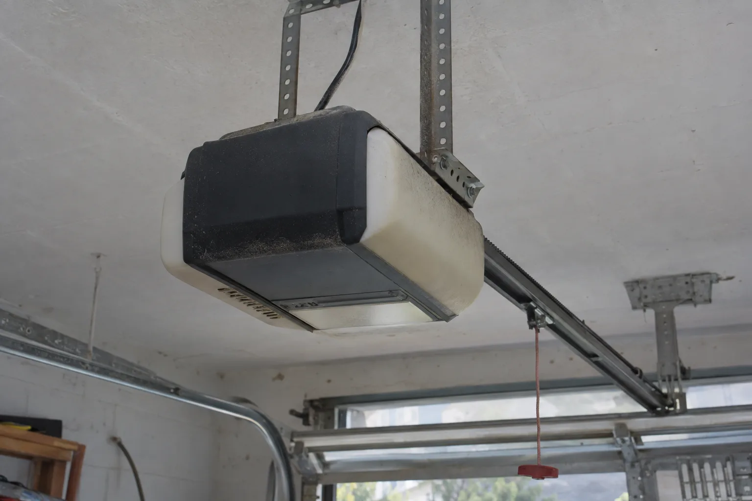 A garage door opener motor unit mounted on the ceiling of a residential garage, with a drive rail extending toward the garage door and daylight visible through the open bay in the background