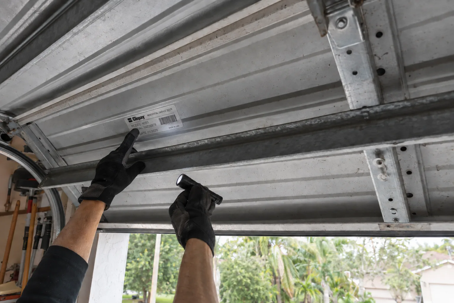 A technician's gloved hands holding a flashlight while inspecting a model identification label on the interior surface of a raised garage door panel inside a residential garage