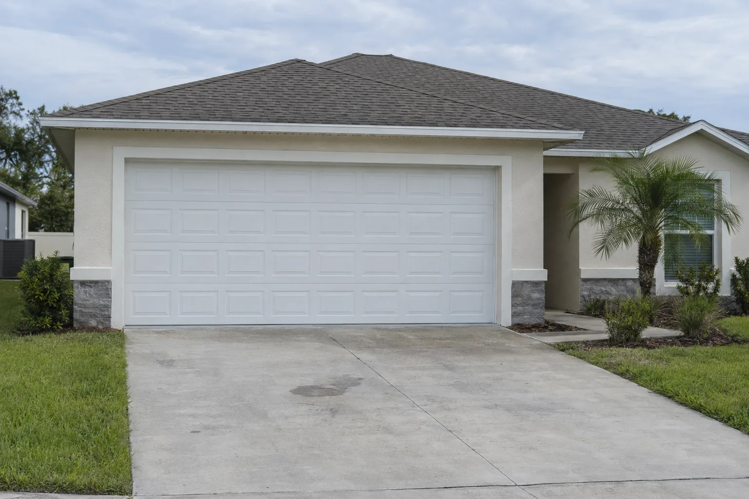 A finished white raised-panel sectional garage door on a newly built single-story stucco home in a Florida suburb, surrounded by a well-kept lawn and a small palm tree under bright daylight