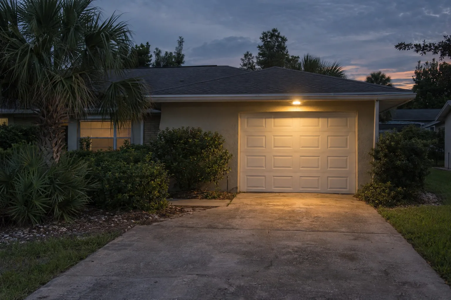 A closed residential garage door on an Orlando ranch-style home photographed at dusk, with a driveway light glowing warmly against a deep blue evening sky and palmetto shrubs along the facade