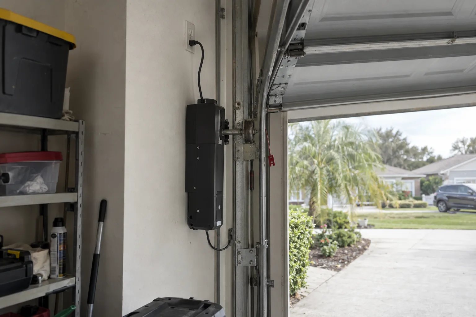 A wall-mount jackshaft garage door opener installed on the interior wall beside a residential garage door track, with wiring routed to a nearby outlet and the garage door visible in the background