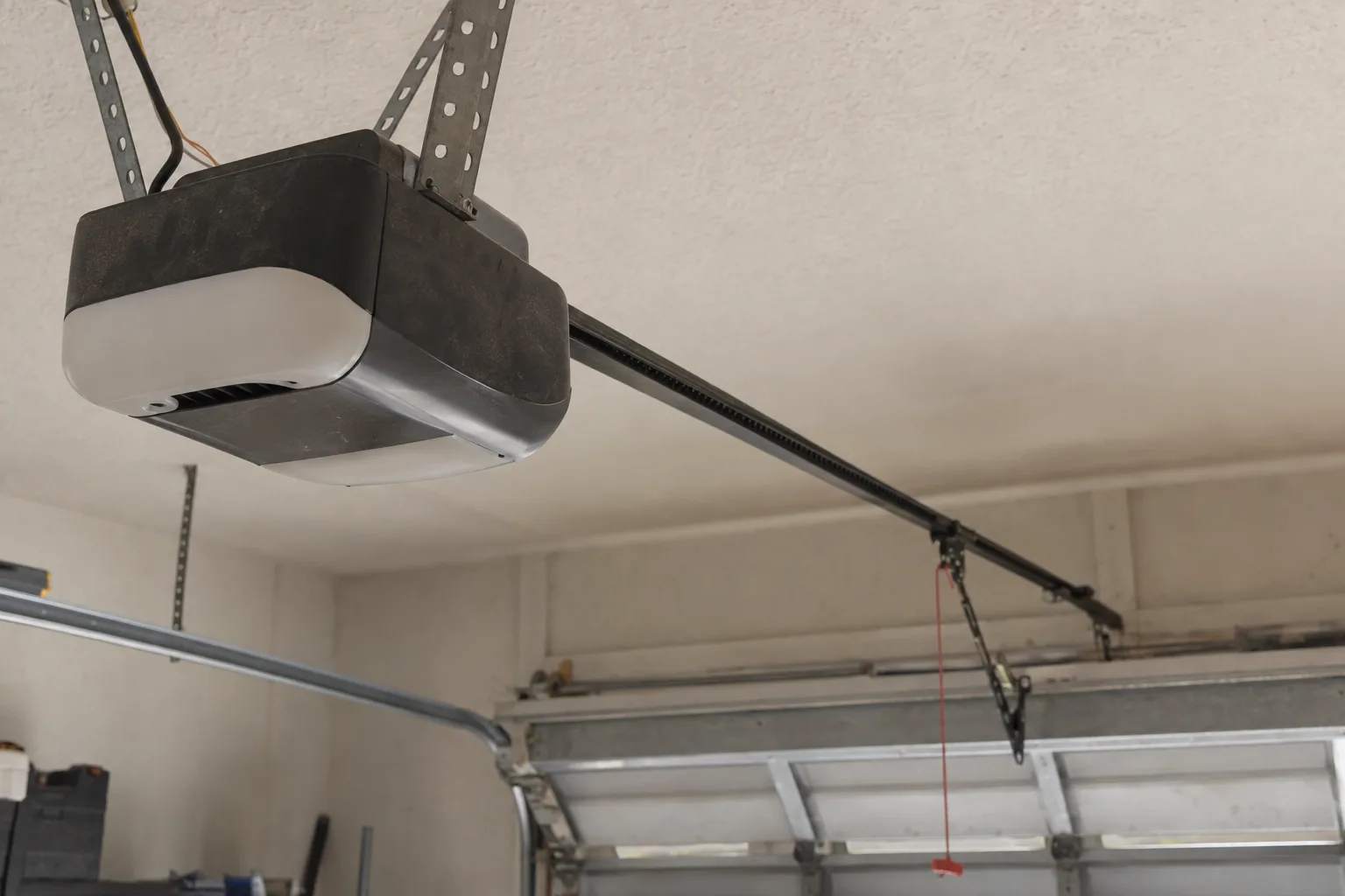 A belt-drive garage door opener and its rail mounted on the ceiling of a residential garage, viewed from below at a low angle showing the drive belt mechanism