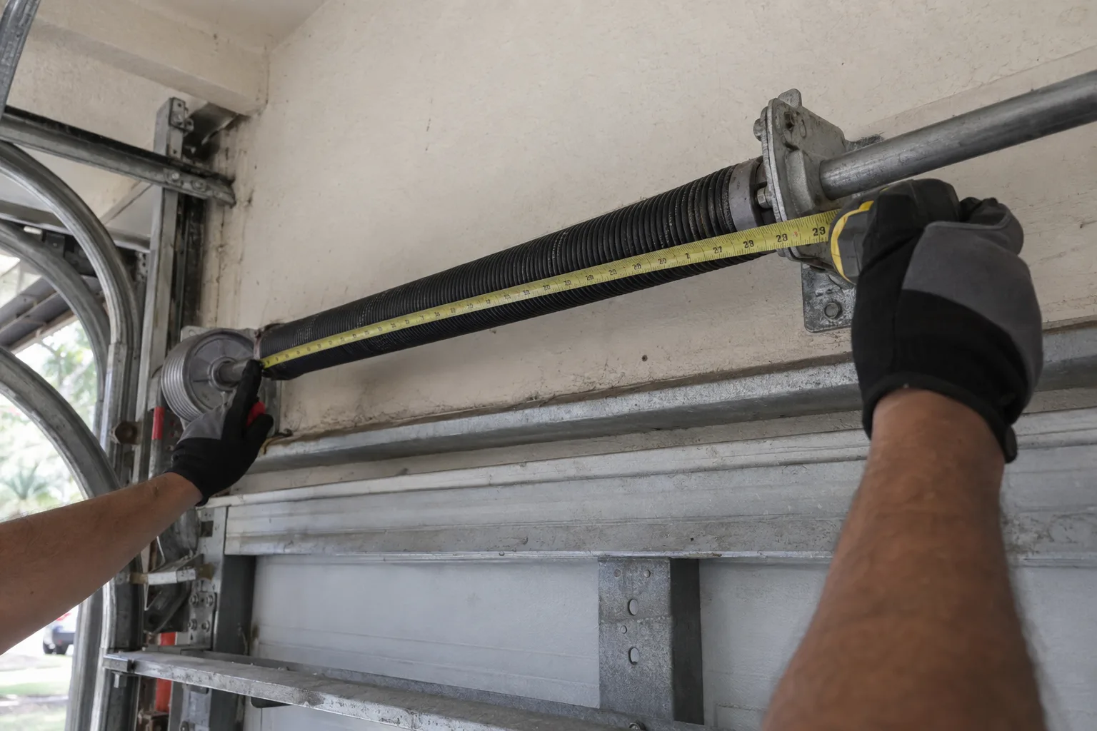 Gloved technician hands measuring a torsion spring mounted on the header bar above a garage door inside a Florida residential garage, with natural daylight illuminating the workspace