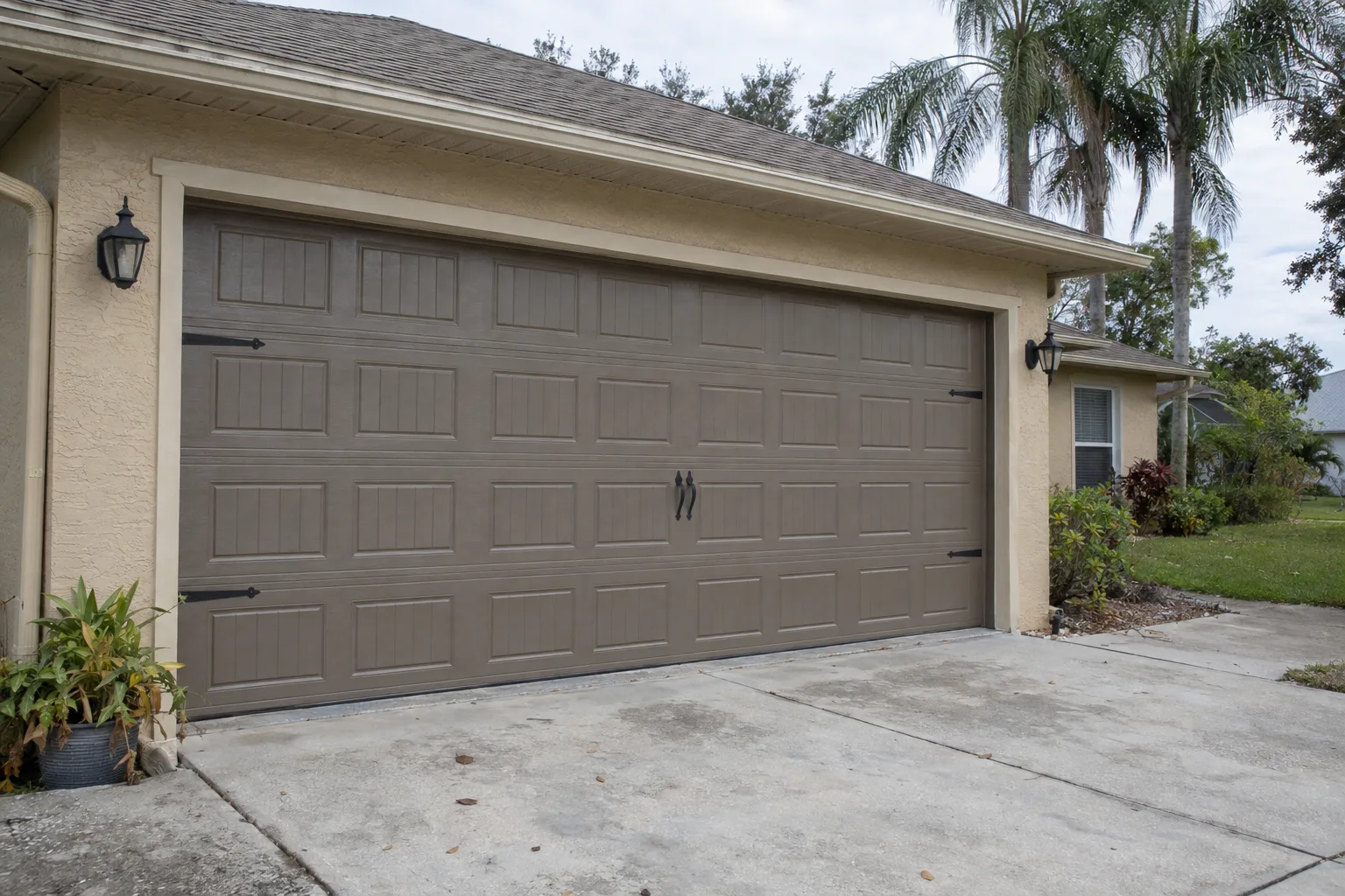 A freshly installed carriage-style garage door with raised panels and decorative hardware on a beige stucco single-story ranch home in a sunny Florida neighborhood, with palm trees visible in the background and a concrete driveway in the foreground