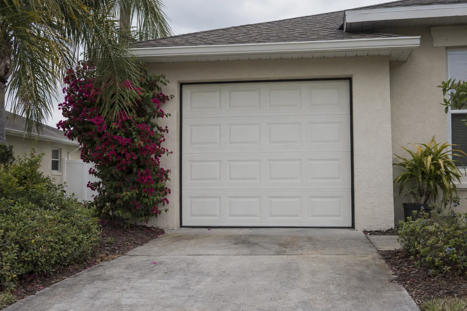 Exterior view of a closed insulated garage door on a Florida stucco home with new black rubber side seals along the door jambs and a bottom seal against the driveway, surrounded by tropical landscaping