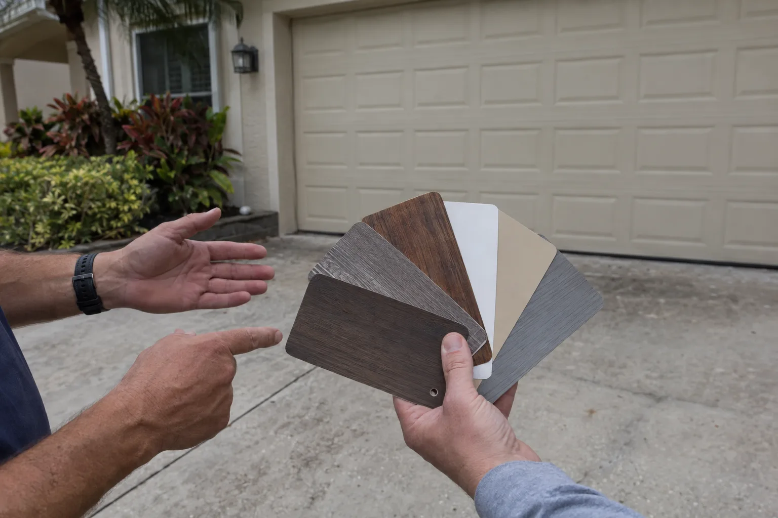 A fan of garage door color and material sample swatches held out in a residential driveway, with a closed garage door softly visible in the background and two sets of hands examining the options