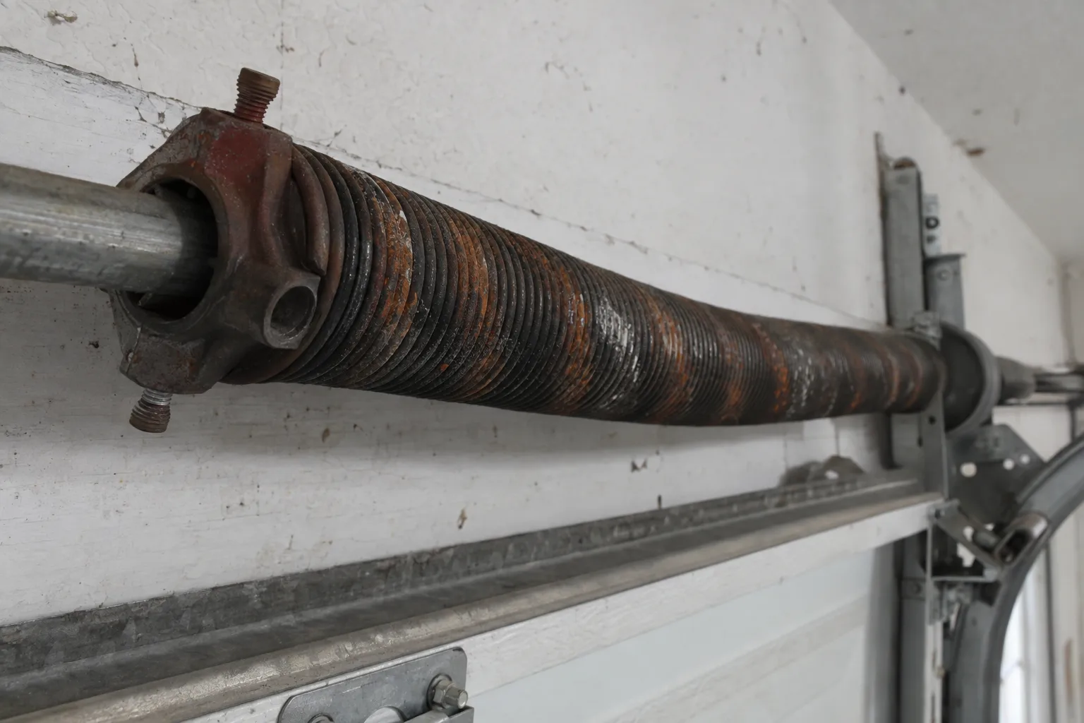 Close-up of a garage door torsion spring mounted above the door on its horizontal shaft, showing patches of orange-brown surface rust and mineral deposits along the steel coils