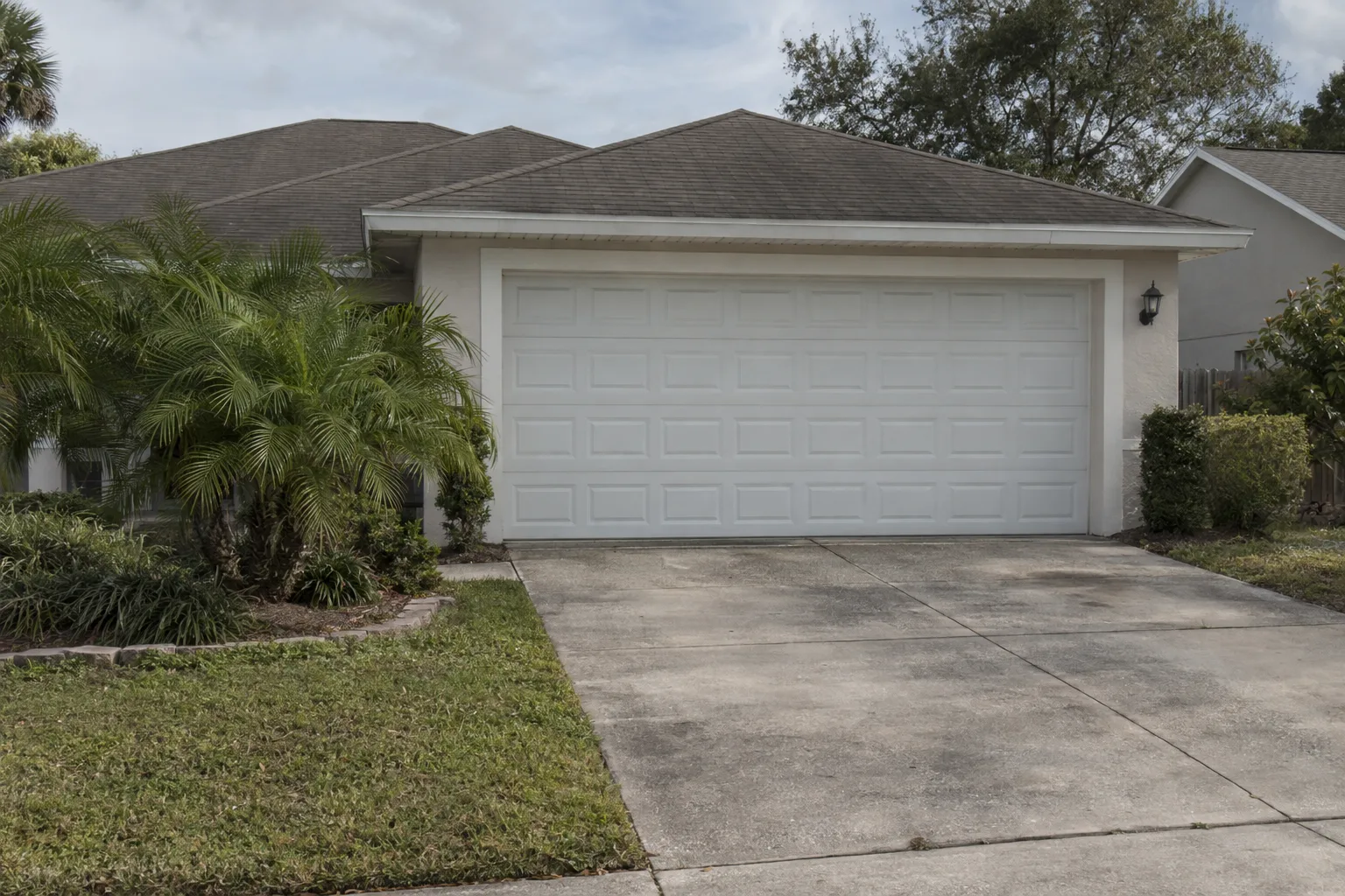 A modern white insulated steel garage door on a single-story ranch-style home in a sunny Florida suburb, with palm shrubs lining the concrete driveway and a blue sky overhead