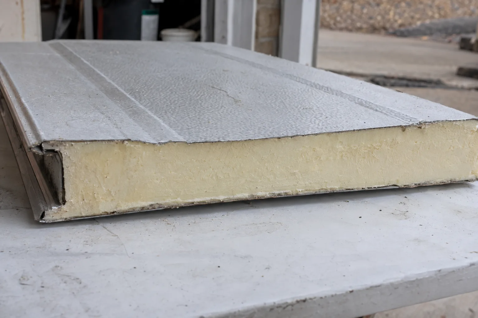 A cross-sectioned garage door panel lying flat, revealing three distinct layers: an outer steel skin, a thick rigid foam insulation core, and an inner steel backing layer, photographed under soft studio lighting